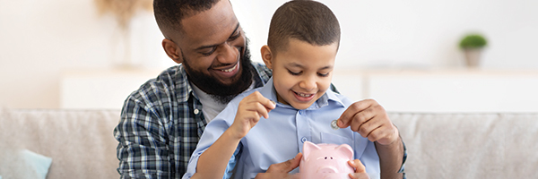 Black man holding his young son on his lap as the boy places coins in a pink piggy bank. They are smiling.