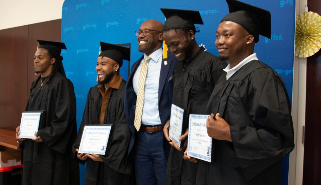 4 young Black men in graduate caps and gowns, holding certificates of achievement and smiling. Goodwill CEO Richmond Vincent stands with them in the center.
